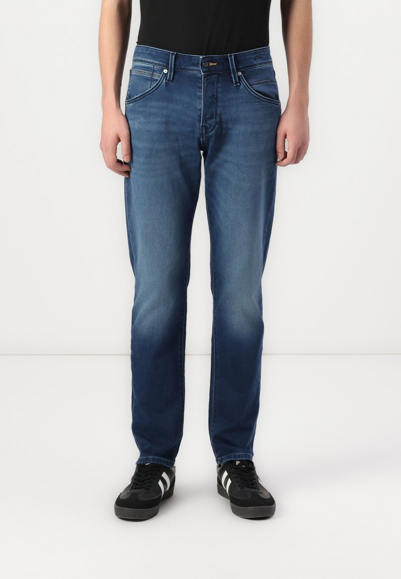 Jean en denim bleu avec une coupe fuselée, doté de poches avant, d'une fermeture éclair et d'un léger délavage. Porté avec des baskets noires et une chemise noire.