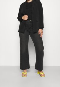 Blazer nero sopra una camicia nera, abbinati a jeans neri a vita alta e gamba larga e sandali con tacco gialli. Elementi di design semplici e moderni.