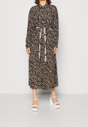 Czarna sukienka z długim rękawem w kwiatowy wzór, z kołnierzykiem, taliowana, z paskiem z białym napisem; noszona z białymi sandałami na platformie.