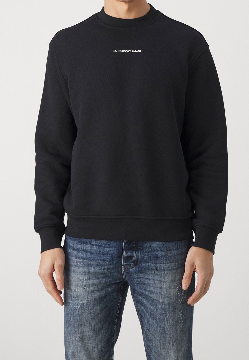 Musta puuvillainen collegepaita pyöreällä pääntiellä ja resoreilla hihoissa. Edessä pieni valkoinen "EMPORIO ARMANI" -logo. Yhdistetty sinisiin farkkuihin.