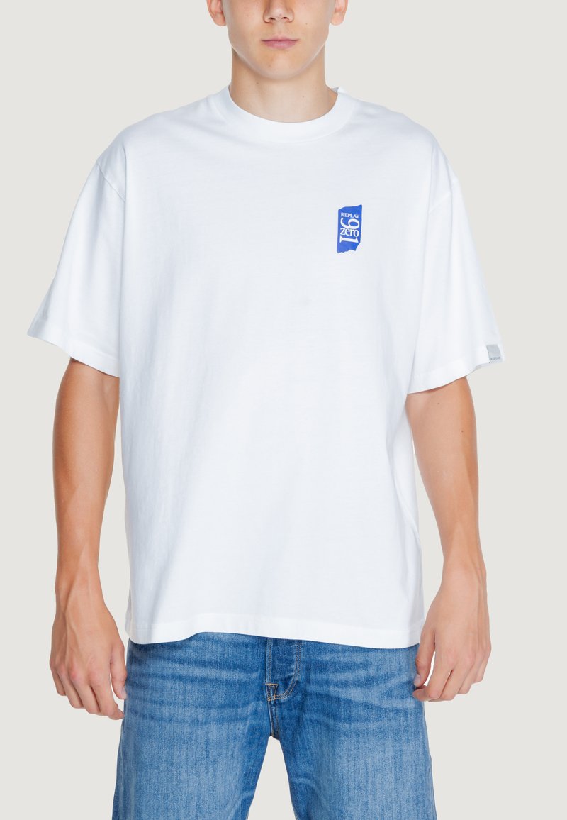 Camiseta de algodón blanca con un parche de logo azul en el pecho izquierdo, mangas cortas y un ajuste relajado. Se lleva con jeans de mezclilla azules.