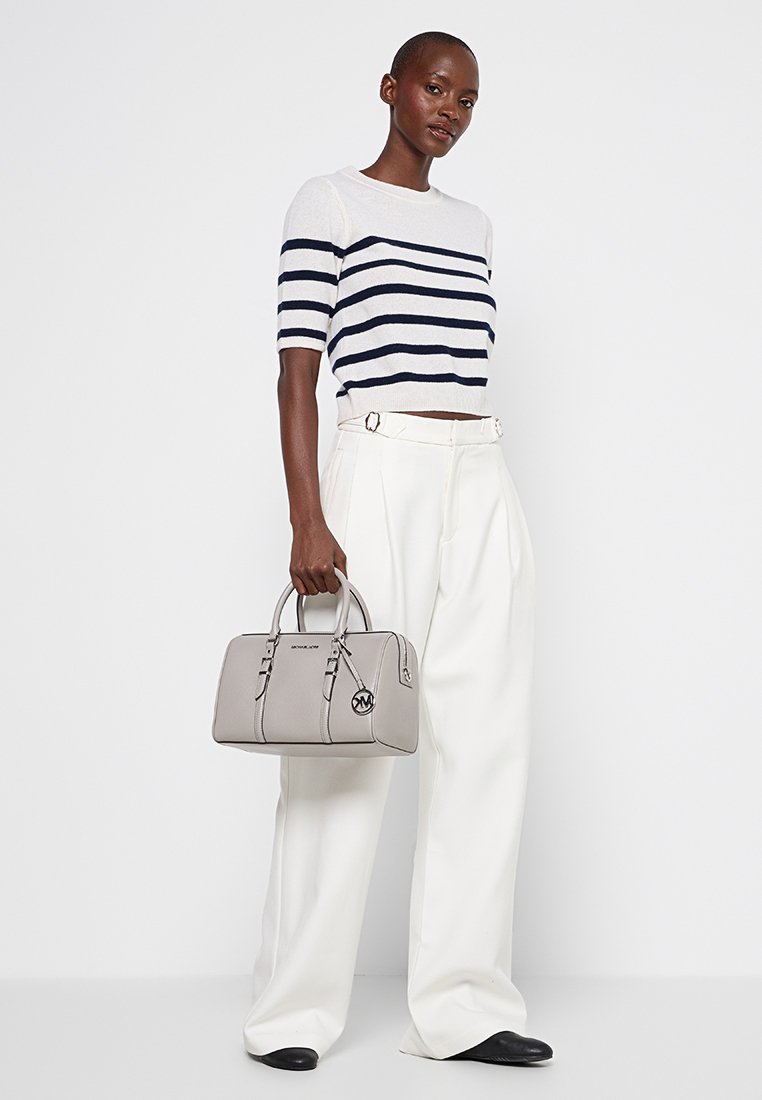 Sac à main en cuir gris au design structuré, avec deux anses supérieures et un logo extérieur. Le modèle porte un pull à rayures à manches courtes et un pantalon blanc.