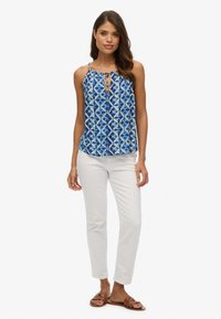 Camiseta sin mangas de color azul con diseños geométricos, tejido ligero, tirantes ajustables, combinada con pantalones blancos ajustados y sandalias marrones.