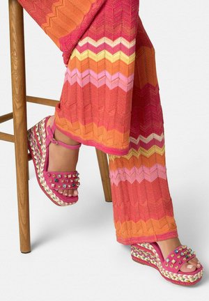 Gambe in pantaloni a maglia a spina di pesce arancioni, rosa e gialli sedute su uno sgabello di legno, indossando sandali rosa con zeppa borchiata e cinturini alla caviglia.