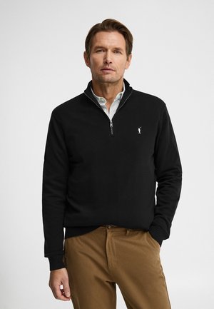 Uomo che indossa un maglione nero con chiusura a mezza cerniera sopra una camicia bianca a righe, con pantaloni marroni, in piedi su uno sfondo chiaro e semplice.