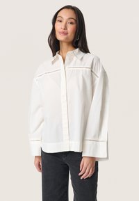 Witte katoenen overhemd met knoopjes en lange mouwen, met uitsparingen op de schouders; gecombineerd met donkere jeans voor contrast.