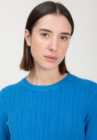 Suéter azul de punto con patrones verticales texturizados y cuello redondo acanalado, que presenta pequeñas perforaciones en toda la tela.