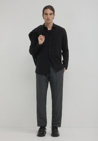 Chemise noire à manches longues, pantalon rayé gris et chaussures noires. Une personne se tient avec un pull drapé sur l'épaule.