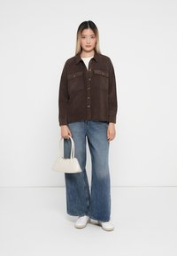 Braune Cordjacke mit zwei Brusttaschen, Knopfverschluss, lockerer Schnitt, kombiniert mit hellblauen weiten Jeans und einer weißen Handtasche.