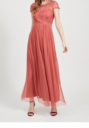 Femme portant une robe longue corail rose avec corsage en dentelle, manches courtes et jupe plissée en tulle, associée à des talons ouverts bordeaux.