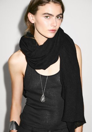 Black scarf made of soft fabric, draped around the neck. Model wears a ribbed black tank top and a silver pendant necklace.