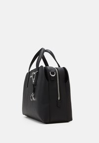 Sac à main structuré noir avec doubles poignées, fermeture éclair argentée sur le côté et breloque logo "ck" argentée sur fond blanc.