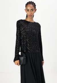 Must mustreeritud must pikkade varrukatega topp ümmarguse kaelusega, koos voolava musta seelikuga ja väikese musta käekotiga. Sile tekstuur, läikivad embellishments.