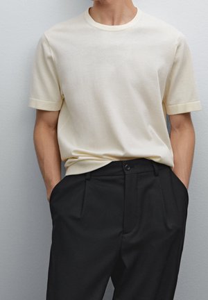 T-shirt beige clair à manches courtes et col rond, associée à un pantalon noir avec plis et poches latérales.