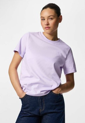 Ung person med mørkt hår bundet tilbage iført en lys lilla T-shirt og mørkeblå jeans med hænderne i lommerne, ensfarvet hvid baggrund.