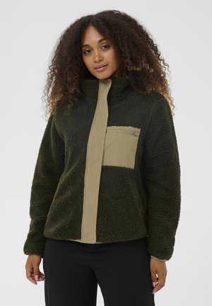 Frau mit lockigem Haar, die eine dunkelgrüne Fleecejacke mit einer hellbeigen, mit Reißverschluss versehenen Brusttasche und Knopfleiste trägt, steht vor einem einfarbigen Hintergrund.