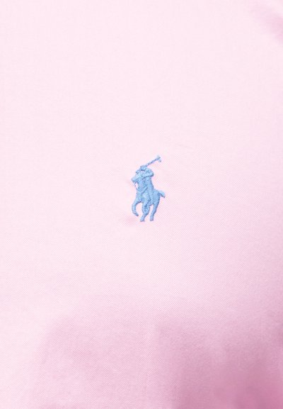 Ανοιχτό ροζ ύφασμα με μικρό ανοιχτό μπλε κεντημένο παίκτη πόλο πάνω σε άλογο που κρατά ανυψωμένο μαλέτι.