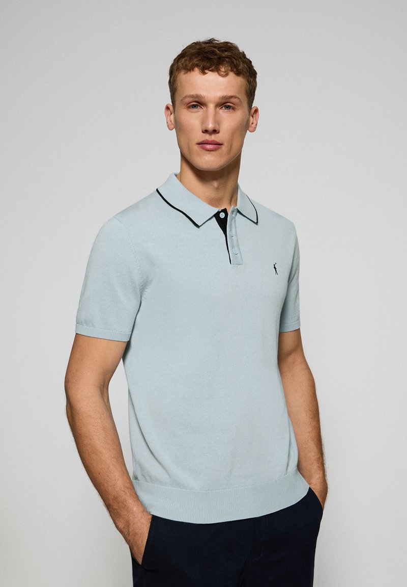 Polo de manga corta azul claro con cuello negro y tapeta de tres botones, que presenta un pequeño logo en el lado izquierdo.