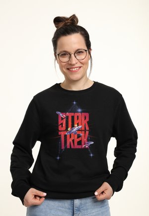 Kvinde med briller og håroppsætning smilende, iført sort sweatshirt med rød "Star Trek"-tekst og rumskibs grafik, holder i kanten.