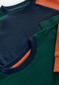 Kaksi crewneck-tyylisiä t-paitaa, tummansinisestä ja vihreästä ribbid-kankaasta, oranssilla yksityiskohdalla olkapäässä, joissa on teksturoitu kuvio ja kontrastivärit.