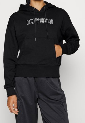 Osoba ubrana w czarną bluzę DKNY SPORT z kapturem i kieszenią kangurką oraz ciemnoszare spodnie cargo, z ręką spoczywającą w kieszeni.