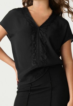 Woman wearing black short-sleeve top with V-neck and ruffled lace trim down the front, paired with high-waisted black skirt.