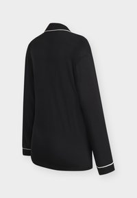 Chemise noire à manches longues avec passepoil blanc sur le col et les poignets, vue de dos sur un fond gris uni.