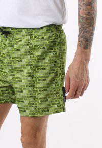 Pantaloni da bagno verdi con stampa totale dei loghi "Emporio Armani" e "EA7", dotati di vita con cordoncino e dettagli con tasche laterali.