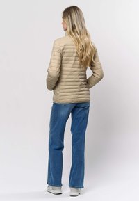 Gaiši bēša šūta jaka ar augstu apkakli, kombinēta ar ziliem džinsiem un baltiem sporta apaviem. Jaka ir ar plūstošu, pieguļošu formu.