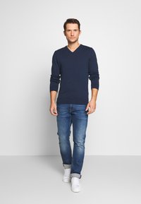 Pulover marin cu decolteu în formă de V din material moale, cu mâneci lungi, ajustat la talie, asortat cu blugi albaștri și adidași albi. Design minimalist, fără modele.