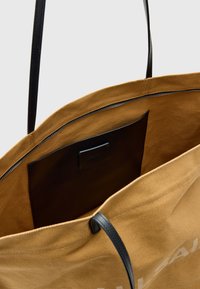 Beige canvas draagtas met zwarte leren handvaten en accenten. De binnenkant heeft een zak van zwart leer. Witte tekst is gedeeltelijk zichtbaar aan de buitenkant.