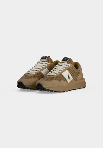 Paire de baskets en mesh et daim marron clair avec lacets blancs et panneau latéral avec un logo de cheval foncé sur fond blanc.