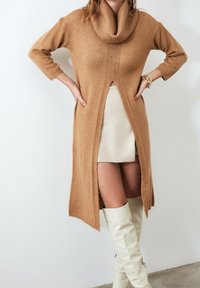 Robe pull en tricot couleur chameau avec un col bénitier, des fentes latérales et des détails côtelés, associée à une jupe claire et des bottes blanches montant jusqu'aux genoux.