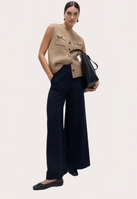 Beige ärmelloses Strickoberteil mit zwei Brusttaschen, dunkelblaue weite Hosen, schwarze Einkaufstasche und passende flache Schuhe. Glatte Texturen überall.