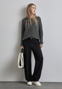 Maglione grigio a maglia con texture a coste, pantaloni neri larghi e una borsa a righe tenuta in una mano; le sneakers bianche completano il look.