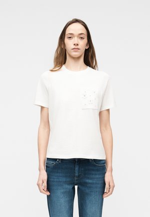 Kvinde iført en hvid kortærmet skjorte med en dekoreret lomme og blå jeans, stående foran en ensfarvet hvid baggrund.