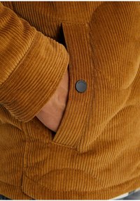 Main dans la poche d'une veste en velours côtelé marron avec un bouton-pression noir visible et des rayures en tissu texturé.