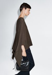 Brauner Strick-Poncho mit lockerem, drapiertem Design. Kombiniert mit schwarzen Hosen und einer kleinen schwarzen Handtasche. Glatte Textur und fließende Form.