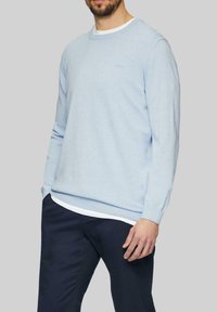 Hellblauer Pullover aus weichem Material, mit Rundhalsausschnitt und gerippten Bündchen. Über einem weißen Hemd getragen, kombiniert mit dunklen Marineblauen Hosen.