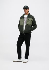 Olive green fleece jacket with zippered pocket, matching panel, black pants, and white sneakers. Worn with a black cap, featuring a logo.
