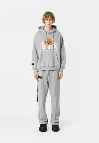 Sudadera gris claro con un gráfico de un perro de peluche, con un corte amplio, bolsillo tipo canguro, combinada con pantalones de chándal grises a juego y zapatos negros con textura.