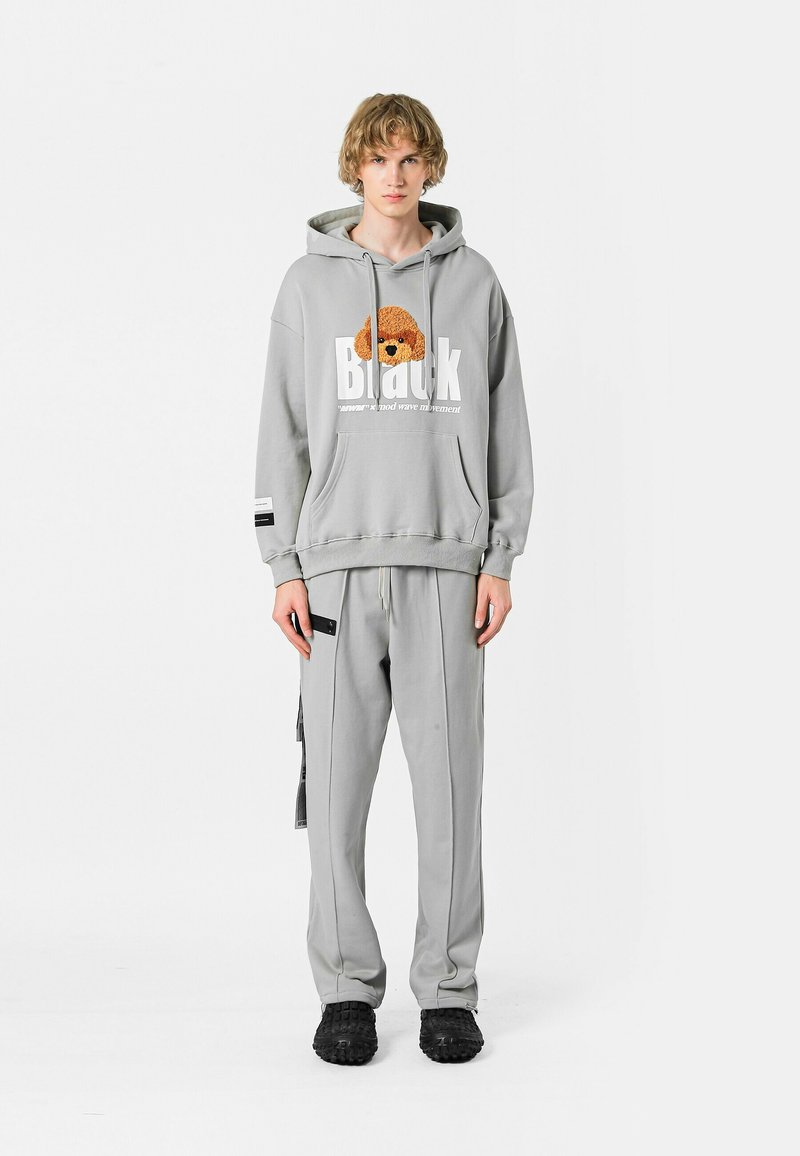 Sudadera gris claro con un gráfico de un perro de peluche, con un corte amplio, bolsillo tipo canguro, combinada con pantalones de chándal grises a juego y zapatos negros con textura.