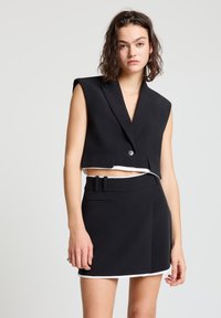 Schwarze, kurz geschnittene Weste mit tiefem V-Ausschnitt und übergroßem Schulterdesign, kombiniert mit einem hoch taillierten schwarzen Rock mit weißer Einfassung.