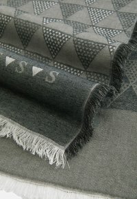 Szary materiał o wzorze z trójkątnych kształtów, z frędzlami na brzegach i kontrastującymi fakturami. Zawiera subtelne białe nadrukowane litery.
