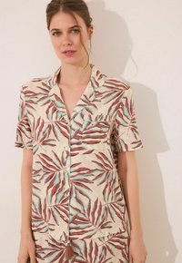 Korte mouwen pyjamashirt in crème met een roestbruine en teal bladerenpatroon. Heeft een zak en een knopenpat met contrasterende bies.