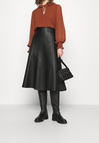 Brun blus med knut vid halsen, svart läder midi-kjol, knähöga svarta stövlar och en kviltad svart handväska. Lätta texturer, enfärgade.