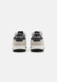 Baskets beiges et grises avec un accent de talon noir, présentant un dessus texturé, un col rembourré et un logo CLAE à l'arrière.
