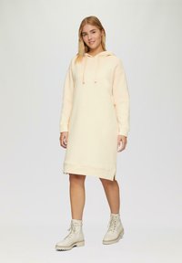 Helle beige Kapuzenkleid mit langen Ärmeln, seitlichen Schlitzen am Saum und einem lockeren Schnitt. Kombiniert mit hellen Ankle-Boots.