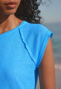Blue cotton t-shirt with a soft, slightly distressed neckline and rolled sleeves. Smooth texture against sunlit skin.