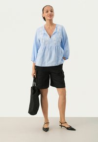 Lyseblå linned blouse med hvid blomsterbroderi, parret med sorte cargo shorts og en vævet sort tote taske, afsluttet med sorte spidse flade sko.